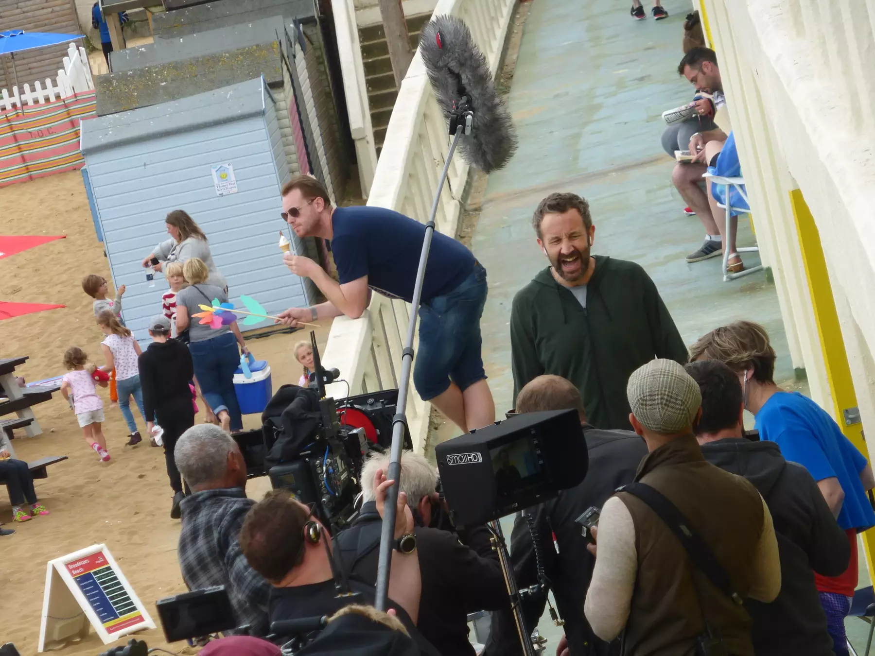 Two ladies and group of children on the beach at the side of a beach hut, Man eating ice cream and holding a windmill leant on white wooden railing. Actor talking to camera with group of film crew behind the camera